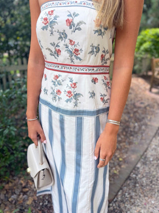 A woman wearing a sleeveless dress with a cream bodice featuring a red and green floral print, a thin red waistband, and a skirt with vertical blue and white stripes. She is holding a small cream handbag in an outdoor garden setting.