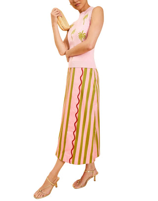 A woman models a sleeveless light pink top with embroidered green palm trees and a midi skirt featuring vertical olive green and pink stripes with a wavy pink center line. She is wearing gold strappy sandals and holding a woven clutch against a solid cream background.