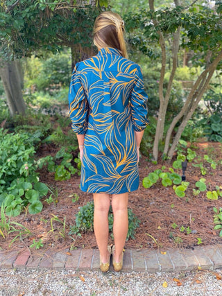 A woman viewed from behind wearing a blue and yellow abstract patterned dress in an outdoor garden setting.