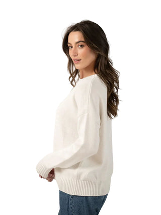 A woman wearing an off-white knit sweater and blue jeans, photographed from the side against a plain light gray studio background.