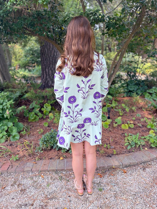 A woman viewed from behind wearing a light sage green long-sleeve tunic dress with a purple floral pattern, standing in a garden setting.