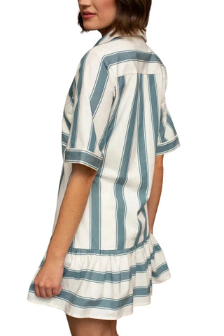 A woman wearing a white short-sleeve shirt dress with vertical blue-gray stripes and a ruffled hem, standing in front of a wooden paneled wall.