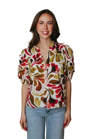 A woman modeling a short-sleeve blouse with a bold abstract floral pattern in red, olive green, mustard yellow, and brown on a cream background, paired with light blue jeans against a solid beige studio backdrop.