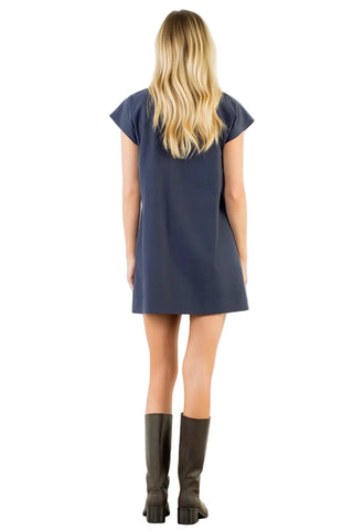 A woman viewed from behind wearing a navy blue short-sleeve shift dress and dark brown knee-high boots, standing against a plain white wall.