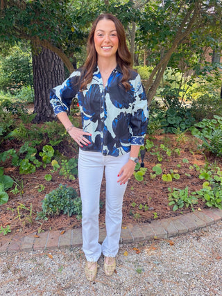 A woman standing outdoors in a garden setting, wearing a blue and black abstract print button-up shirt, white pants, and beige shoes.