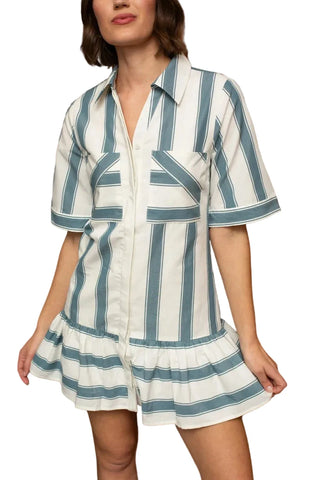 A woman wearing a short-sleeve collared mini dress with vertical blue and white stripes, standing in front of a dark wood-paneled background.