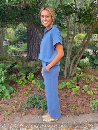 A woman wearing a matching blue two-piece lounge set consisting of a short-sleeve top and wide-leg pants, standing outdoors in a garden setting.
