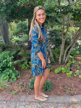 A woman stands outdoors in a garden wearing a blue collared shirt dress with a gold botanical pattern, paired with beige flat shoes.