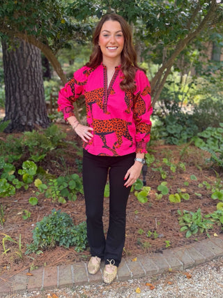 A woman standing outdoors in a garden setting, wearing a bright pink long-sleeve button-up shirt with a bold leopard print pattern, black pants, and beige shoes.