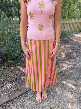 A woman standing outdoors on a gravel path wearing a pink sleeveless knit top with green palm tree prints and a matching pink and olive green striped midi skirt.