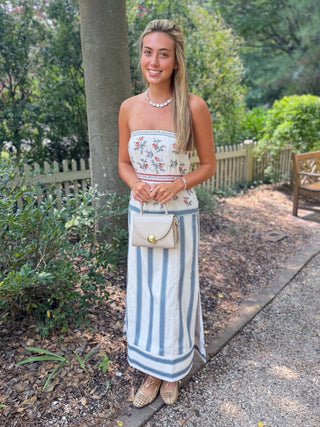 A woman stands outdoors on a gravel path in a garden, wearing a strapless tube dress with a floral top section and blue-and-white vertical stripes on the skirt, holding a small beige handbag.