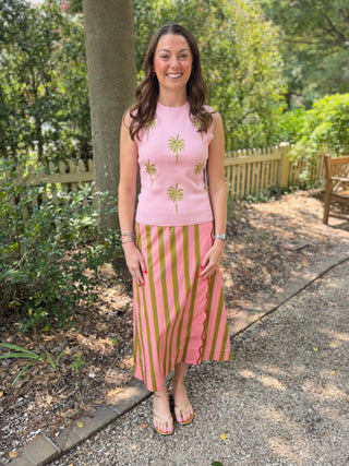 A woman stands on a gravel path in a garden setting, wearing a light pink sleeveless top with green palm tree embroidery and a matching pink and green vertically striped midi skirt.