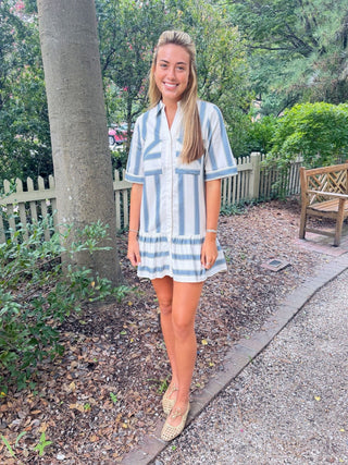 A woman stands outdoors on a gravel path wearing a short-sleeve button-front mini dress with vertical blue and white stripes, paired with beige flat shoes. Trees, a bench, and a picket fence are visible in the background.