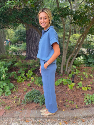 A woman wearing a matching blue two-piece lounge set consisting of a short-sleeve top and wide-leg pants, standing outdoors in a garden setting.