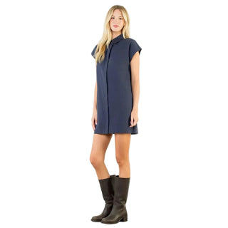 A woman modeling a navy blue short-sleeve collared shirt dress with dark brown knee-high boots, standing against a plain white studio wall.