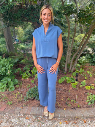 A woman stands outdoors in a garden wearing a matching blue sleeveless top and wide-leg pants set, with a small zipper detail at the neckline.