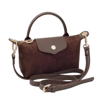 A dark brown suede handbag with a top handle, a flap closure featuring a gold-tone button, and a detachable shoulder strap, styled on a white textured surface with a decorative book in the background.
