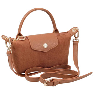 A brown suede-look handbag with a top handle, front flap, and detachable shoulder strap, styled on a white textured surface with books and a curtain in the background.