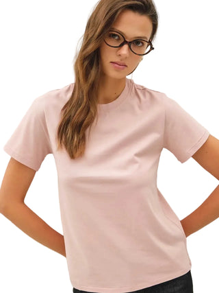 A woman with long brown hair and glasses wearing a light pink crew-neck t-shirt, posing with hands on hips against a plain off-white background.