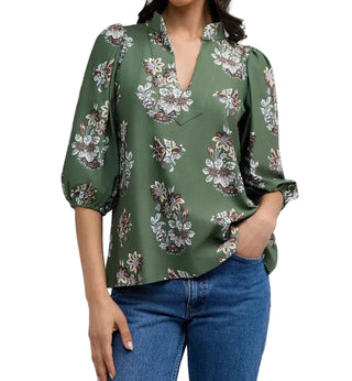 A woman wearing a sage green long-sleeve blouse with a white and brown floral pattern, paired with blue jeans, shown against a plain light gray background.