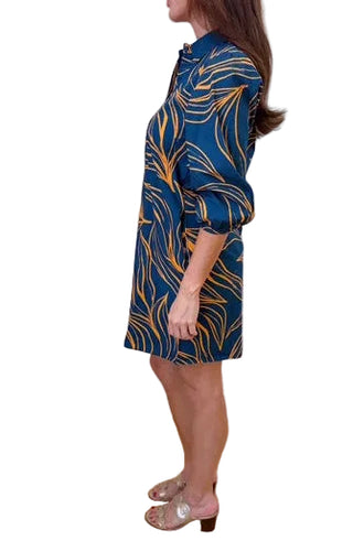A woman stands in profile wearing a blue satin-like shirt dress with a gold abstract leaf pattern, paired with metallic silver sandals, in an indoor room with white walls and a light wood floor.