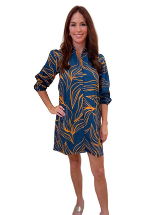 A woman modeling a short-sleeve button-up shirt dress with a blue and mustard yellow tropical leaf pattern, standing on a light wood floor against a white wall.