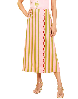 A model wearing a pink midi skirt with vertical olive green stripes and a red wavy center stripe, paired with a pink top and gold heeled sandals, set against a solid cream background.