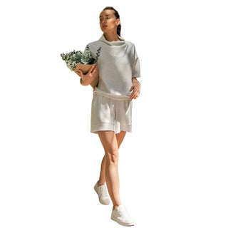 A woman walking outdoors on a brick path wearing a light beige knit cowl-neck tunic top and matching shorts, holding a small potted plant.