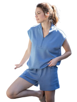 A woman wearing a matching light blue lounge set consisting of a sleeveless quarter-zip top and matching shorts, posing outdoors next to a pool with a blue wall in the background.