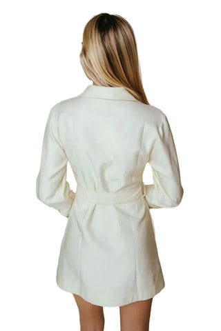 A woman viewed from behind wearing a cream-colored long-sleeve wrap coat or dress with a tie belt, set against a solid beige background.
