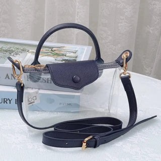 A clear plastic handbag with dark navy blue leather trim, a top handle, a flap closure with a snap button, and a detachable shoulder strap, styled on a white lace-covered surface with books in the background.