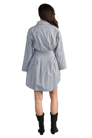 A woman viewed from the back wearing a light blue belted shirt dress with three-quarter sleeves and black ankle boots, set against a solid gray studio background.