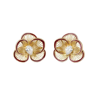 A pair of gold flower-shaped stud earrings with dark red enamel outlines and a clear marquise-cut gemstone in the center, displayed on a light gray textured fabric background.