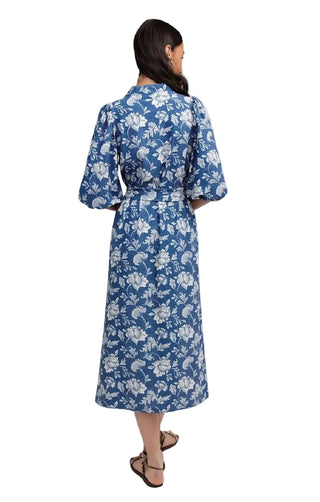 A woman viewed from the back wearing a blue midi dress with a white floral pattern, three-quarter sleeves, and a tie waist, paired with brown strappy sandals against a light gray studio background.