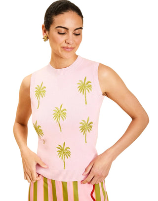 A woman wearing a sleeveless light pink knit sweater with green palm tree motifs, paired with pink and green striped bottoms, against a solid beige background.