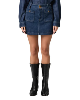 A model from the waist down wearing a dark blue denim mini skirt with front patch pockets, a matching denim long-sleeve top, and black leather knee-high boots, set against a plain light gray studio background.