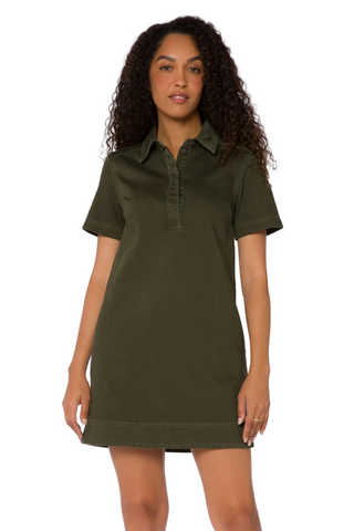A woman with curly dark hair wearing an olive green short-sleeve polo shirt dress, standing against a plain white studio background.