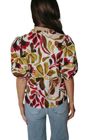 A woman viewed from the back wearing a short-sleeve blouse with a bold floral pattern in red, olive green, brown, and cream, paired with light blue jeans against a plain beige studio background.