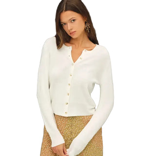 A woman wearing a fitted white long-sleeve cardigan with a split neckline and gold-tone buttons, paired with a patterned skirt, posed against a dark gray studio background.