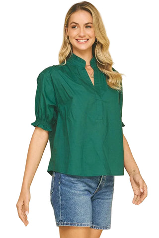 A woman modeling a dark green short-sleeve blouse with a high mock neck and puff sleeves, paired with blue denim shorts, against a solid beige background.